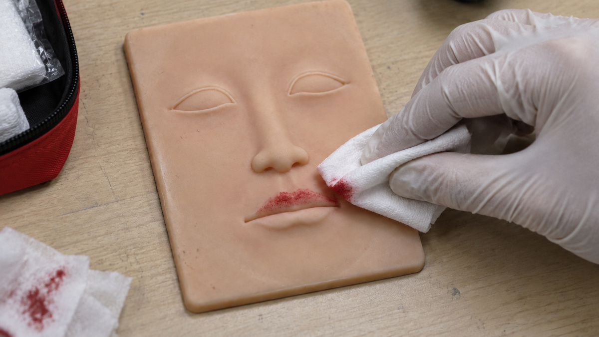 A gloved hand wipes red pigment from the lips of silicone practice skin