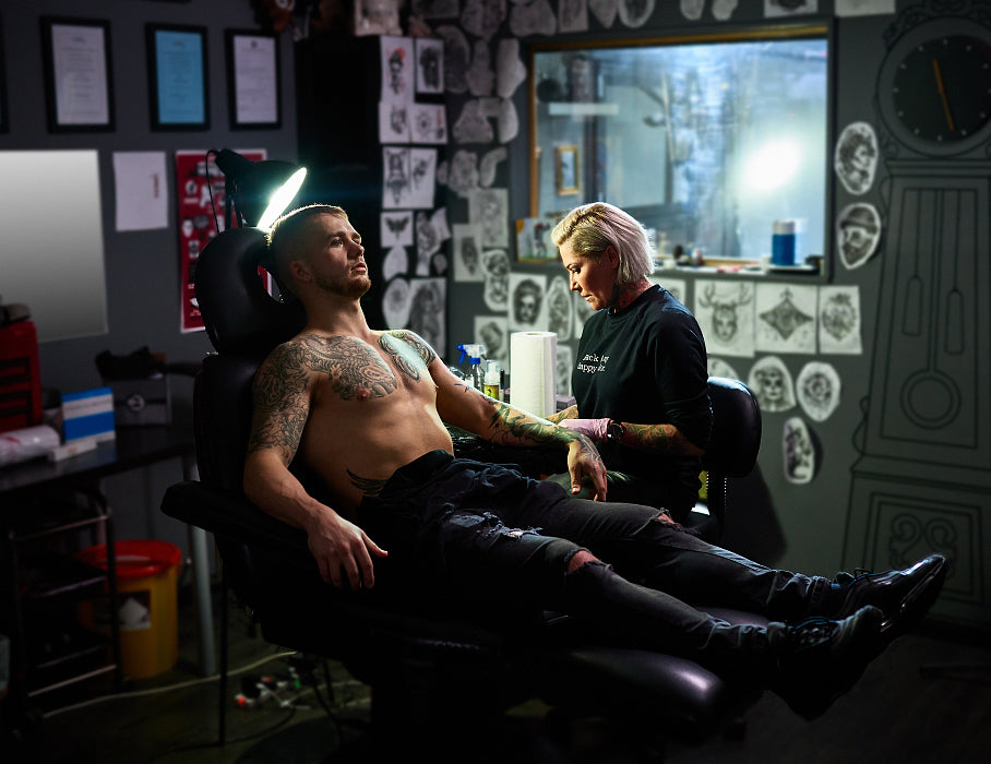 In a tattoo studio, a man with existing tattoos relaxes in a black leather tattoo chair while a female tattoo artist concentrates on tattooing his arm.