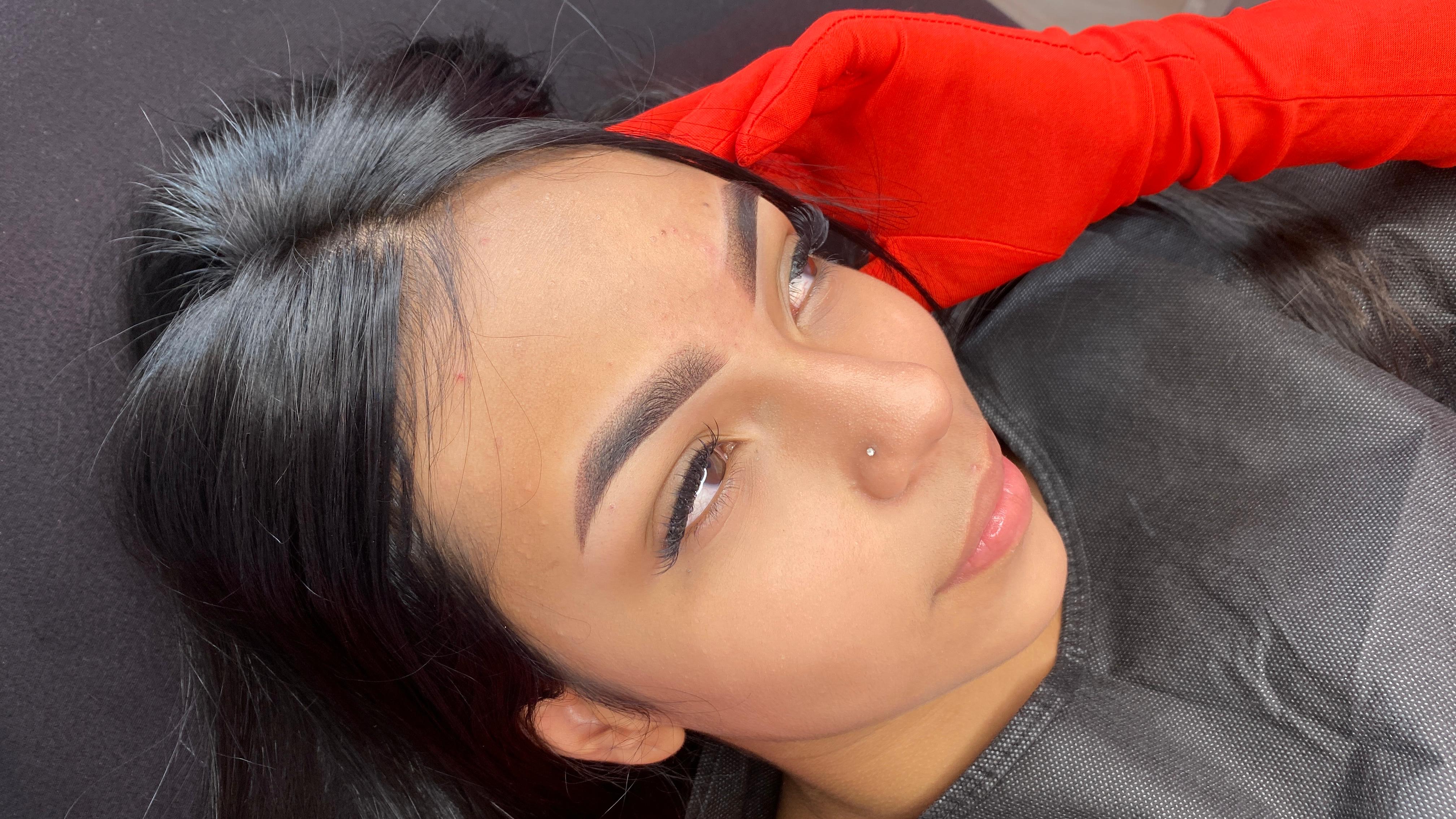 Woman receiving eyebrow microblading treatment at beauty salon, lying down with a red headband protecting her hair, showcasing the eyebrow beauty care process.
