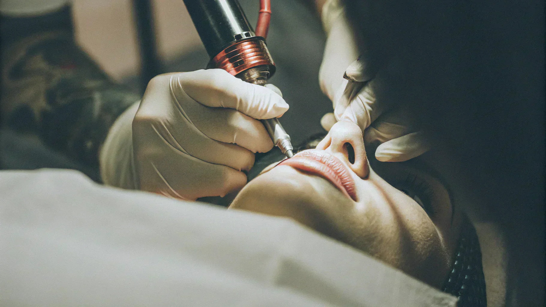 Professional semi-permanent lip makeup procedure in progress, with a beautician wearing gloves using an electric micropigmentation device for precise lip contour design and pigment application.